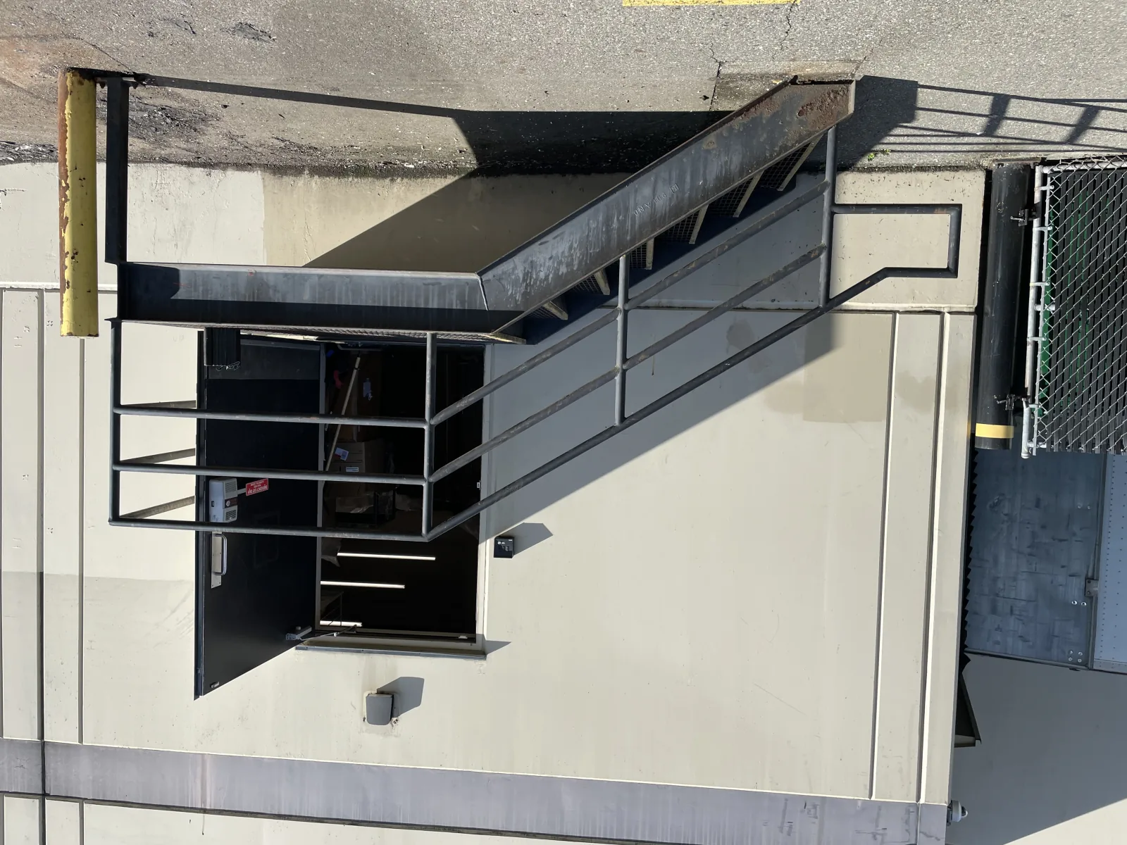 Black-painted exterior platform stair at a Vancouver loading bay with pipe-rail guards, bar grating treads, and a building-mounted landing — a current code-compliant replacement of an older pan-tread stair