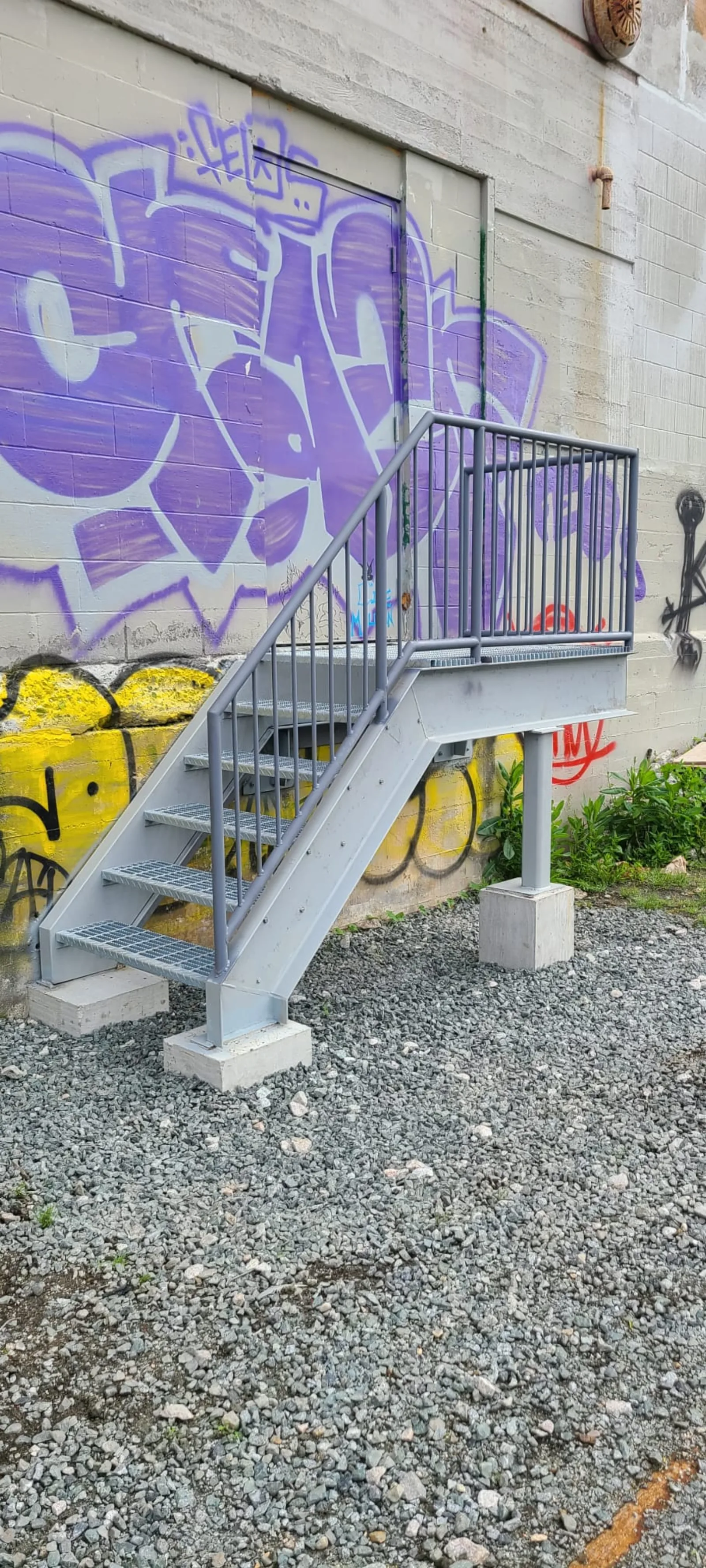 Short four-step galvanized platform stair with bar grating treads, pipe-rail guards, and concrete post bases — a back-entry warehouse replacement at a graffitied light-industrial building